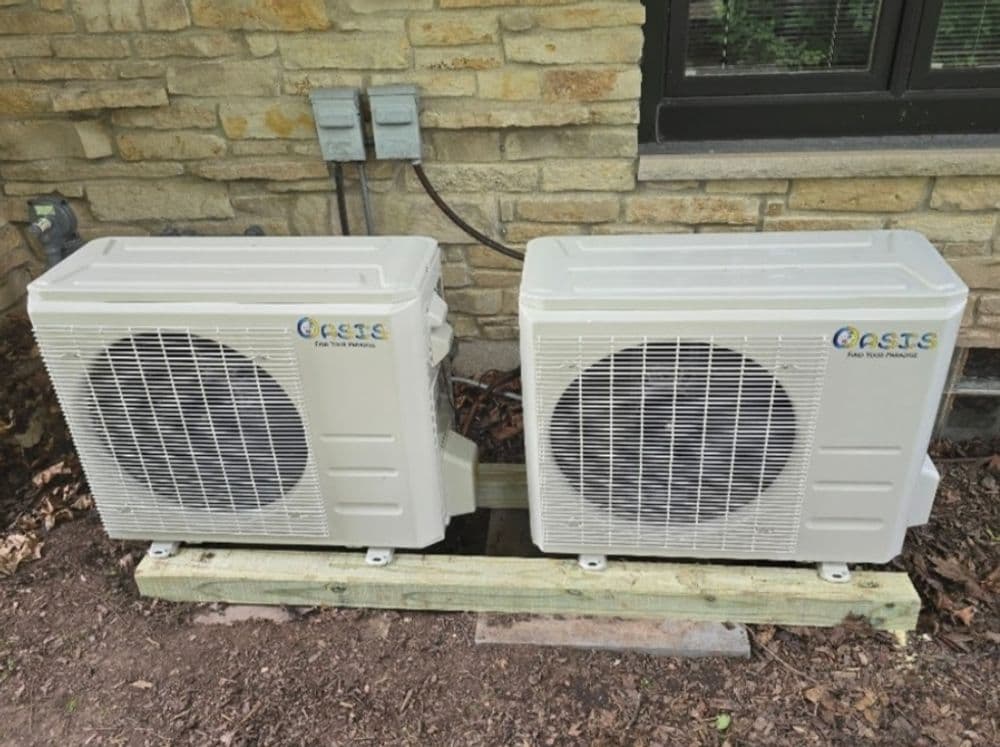 Two outdoor air conditioning units mounted on wooden platform near a stone wall.
