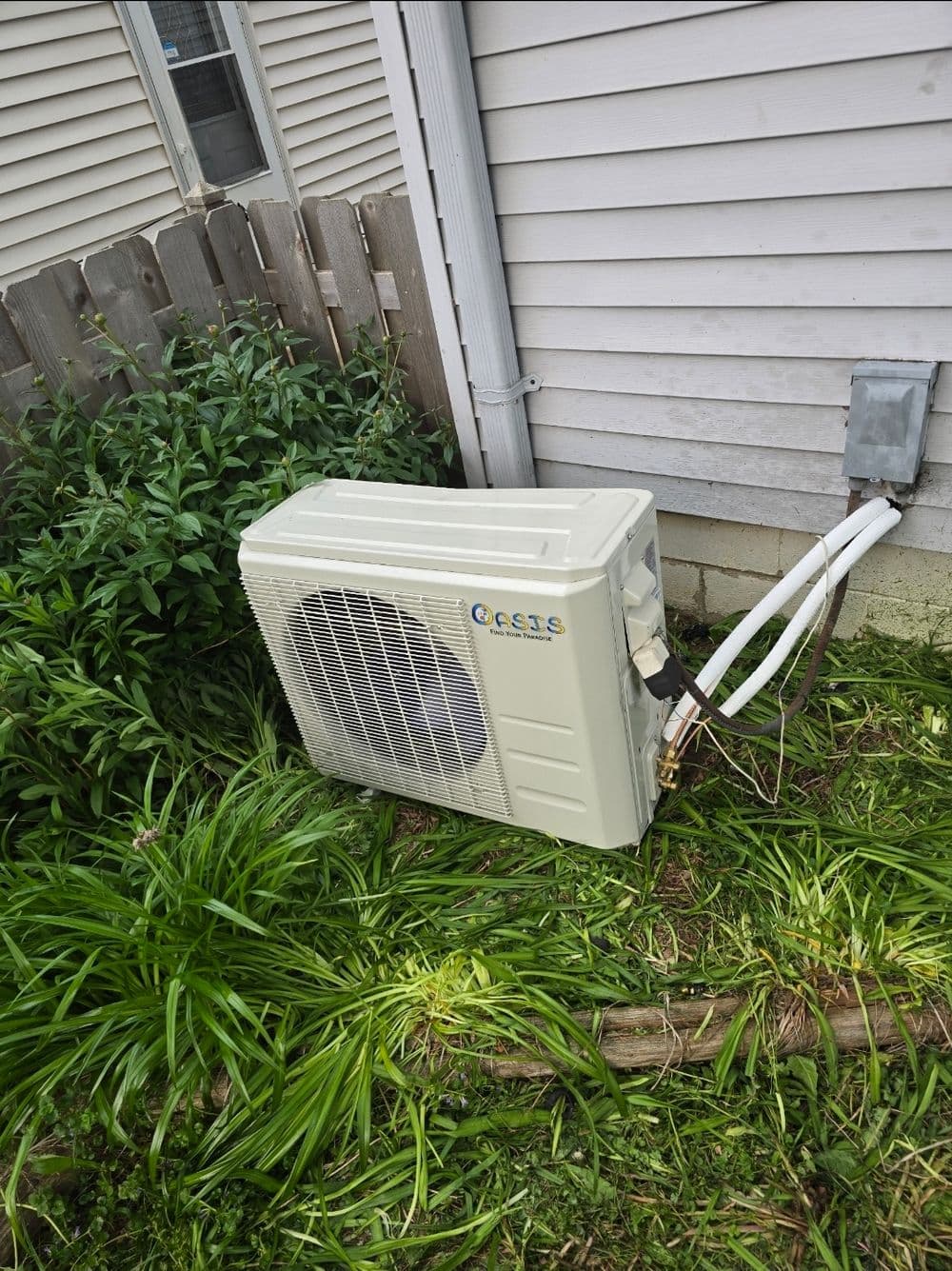 Outdoor air conditioning unit next to a house, surrounded by overgrown grass and plants.
